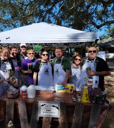 Group of ten people wearing aprons posing behind a decorated table at an outdoor event under a white canopy tent.