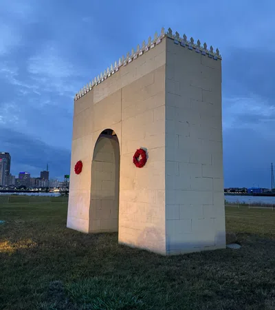 Large white stone monument with red wreaths on a grassy field at dusk with a city skyline in the background