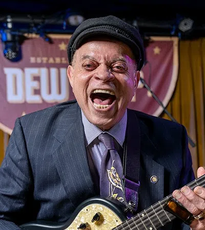 Smiling man in a suit and hat playing electric guitar on a stage with wooden backdrop and lights