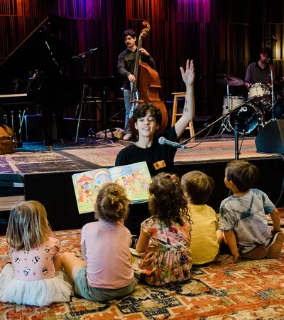 Adult reading a colorful book to children seated on a rug with live musicians performing in the background