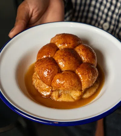 Person holding a white plate with golden brown sticky buns drizzled in syrup inside a rustic setting