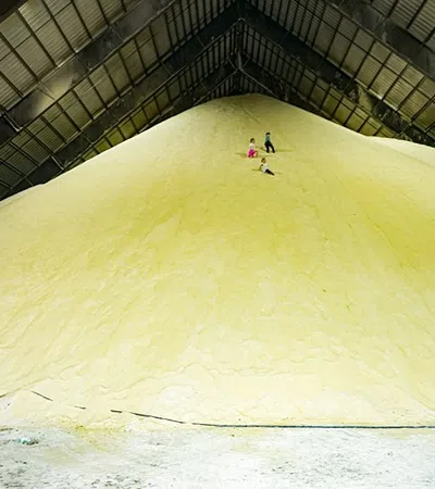Children play on a large yellow sulfur pile inside an industrial warehouse with a woman watching.