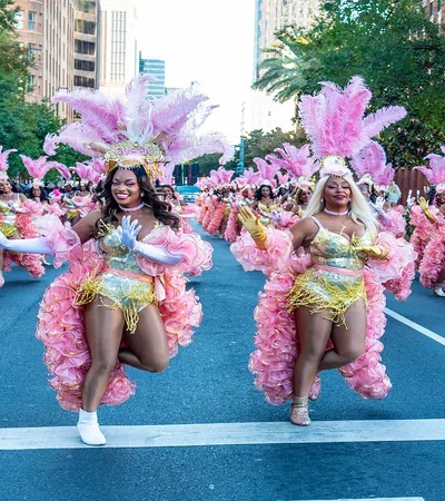 Dancers in pink feathered costumes perform in a vibrant street parade in a city setting, smiling and energetic.