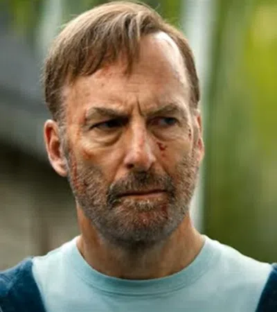Intense bearded man with facial cuts looks serious outdoors with blurred greenery and person in foreground