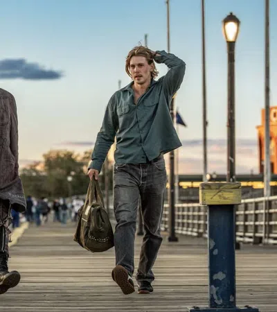 Two young men walking on a wooden pier at sunset, one with a colorful mohawk and punk style, the other casual with a bag.
