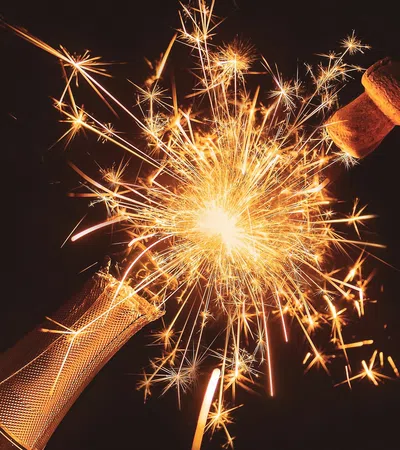 Champagne bottle popping with sparkling fireworks bursting from the neck against a dark background.