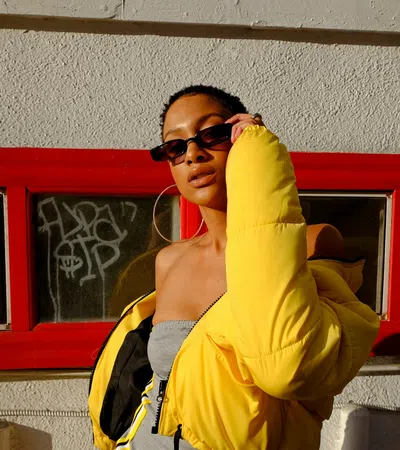 Woman in a bright yellow puffer jacket and sunglasses poses confidently against a textured wall with red window frames.