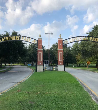 Entrance to Lafreniere Park with brick pillars, black iron archway, and tree-lined roads under blue sky.