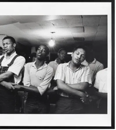 Black and white photo of young men standing with arms linked in a room with exposed ceiling light.
