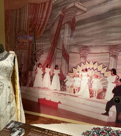 Vintage beaded white gown displayed next to a stage backdrop with women in white dresses during a pageant.