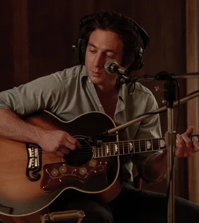 Man wearing headphones playing an acoustic guitar and singing into a microphone in a recording studio.