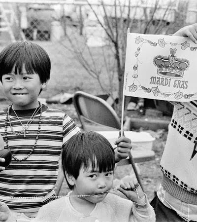 Three children smiling and playing outdoors, one holding a Mardi Gras flag, others wearing necklaces and festive clothes.