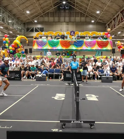 Indoor pickleball doubles match with players poised at the net and a large colorful audience in the background