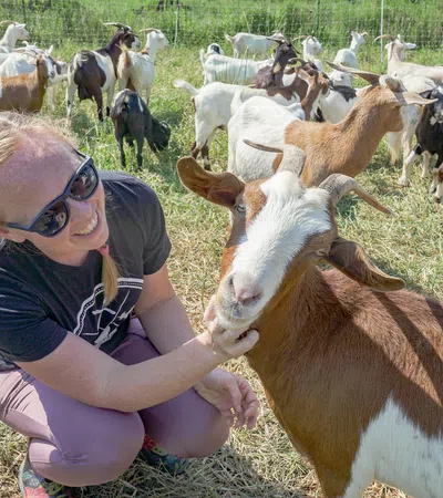 Woman wearing sunglasses smiling and petting a brown and white goat in a field with many goats.