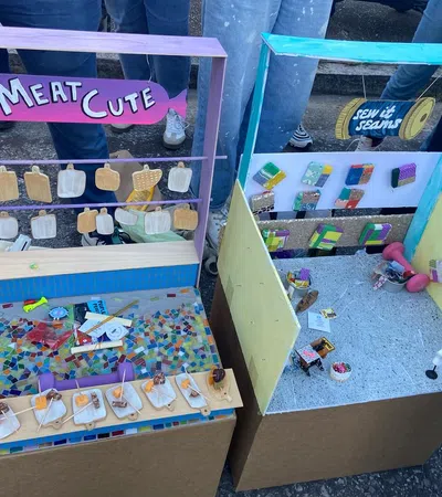 Two colorful miniature market stalls with handmade items labeled Meat Cute and Sew It Seams displayed outdoors with people nearby.