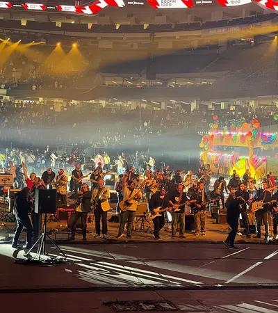 Large group of musicians performing on stage in a stadium with colorful lights and audience in background
