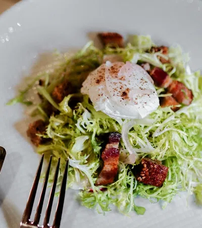 Fresh frisée salad with crispy bacon, croutons, and a poached egg on a white plate with a fork and knife.