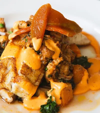 A beautifully plated dish with grilled fish, vegetables, and a rich sauce garnished with a slice of orange.