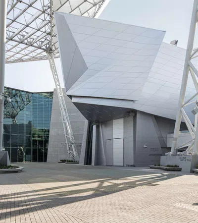 Modern architectural building with angular metal panels and large steel supports casting shadows on a paved courtyard.
