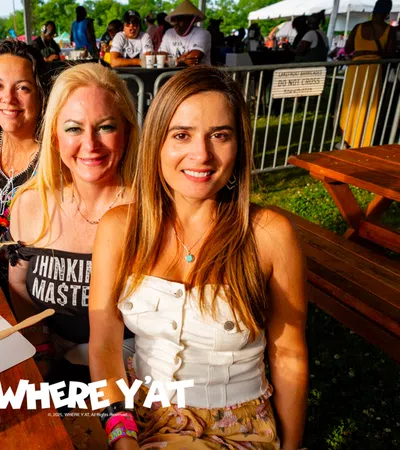 Three smiling women sitting at a wooden picnic table at an outdoor festival with drinks and beverages visible.