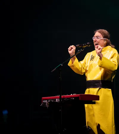 Performer in yellow costume sings passionately at microphone with keyboard and drum set in background on stage