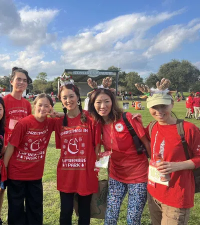 Group of seven people wearing festive red shirts and reindeer antlers posing outdoors at a park event on a sunny day