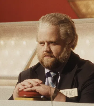 Man with beard and suit pressing a buzzer on a game show set with name tag reading Michael.