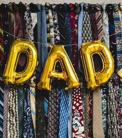 Gold balloons spelling DAD hang among rows of colorful patterned men's neckties on a wall display.