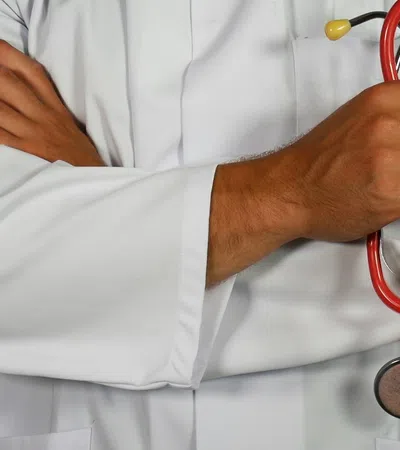 Doctor in white coat holding red stethoscope with arms crossed against light background