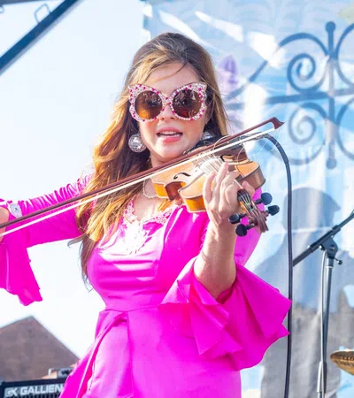 Woman in bright pink dress playing violin on outdoor stage during daytime musical performance.