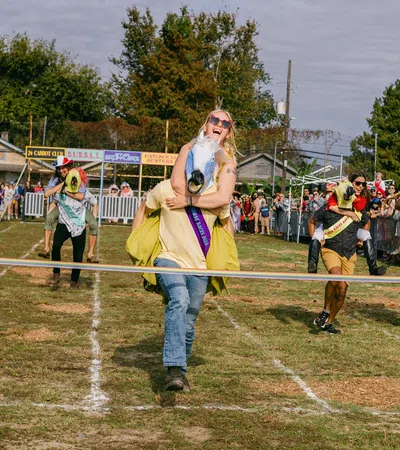 Couples race in a horse costume event on a grass track with spectators cheering behind barriers.