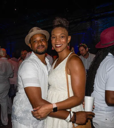 Couple in white outfits smiling and embracing at a lively indoor party with colorful lighting and crowd.