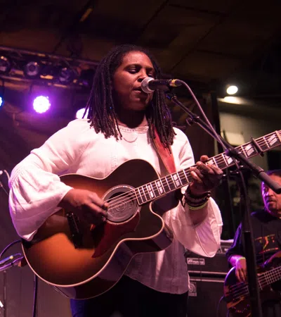 Female musician singing and playing guitar on stage with band at Crescent City Blues festival under purple lights.