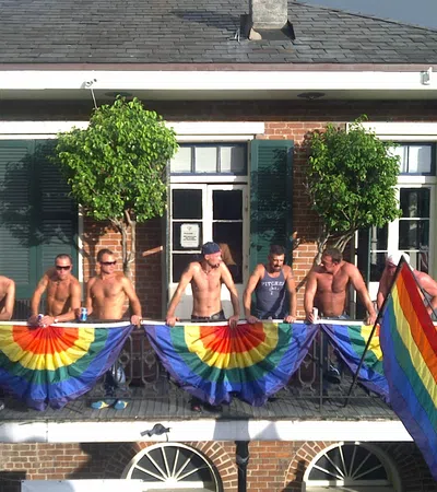 Group of shirtless men holding rainbow flags on a balcony outside a brick building under sunny sky