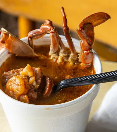 Close-up of a seafood soup with crab legs and shrimp in a white cup with a black plastic spoon.