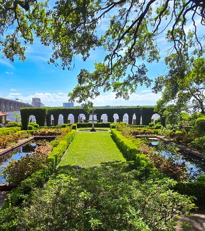 Elegant garden with manicured hedges, reflective pools, stone benches, and arched hedges overlooking a water body.