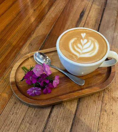 White cup of latte with heart-shaped latte art on a wooden platter with purple flowers and spoon.