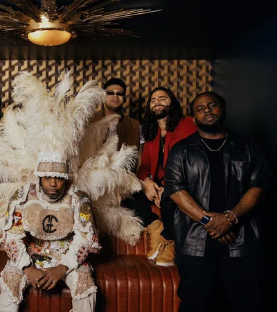 Group of seven men posing indoors with one in an elaborate feathered costume and others in casual and stylish attire