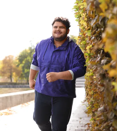 Smiling man in purple jacket jogging outdoors along a wall with autumn foliage in the background.