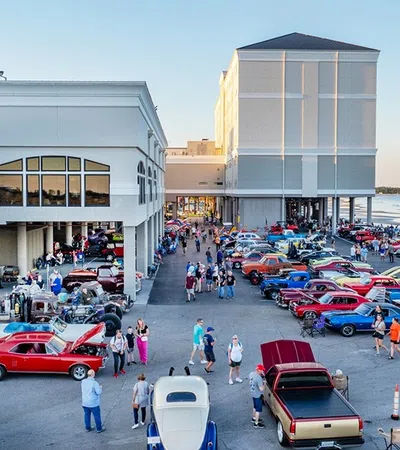 Outdoor classic car show with vintage cars and attendees near waterfront buildings at sunset.