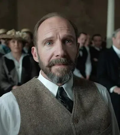 Man with beard and vest sitting attentively in a vintage courtroom with formally dressed audience behind him.
