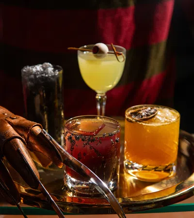 Person in striped sweater and clawed glove serving four colorful cocktails on a round tray at a bar