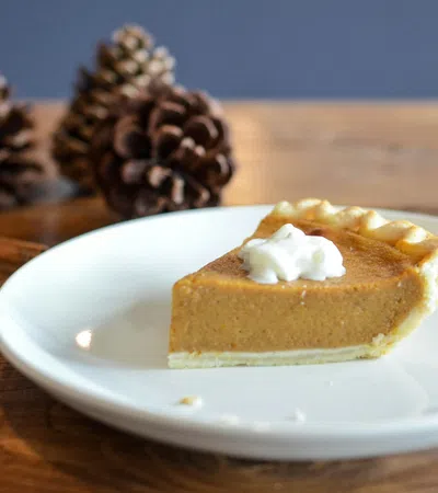 Slice of pumpkin pie with whipped cream on white plate with pine cones on wooden table.