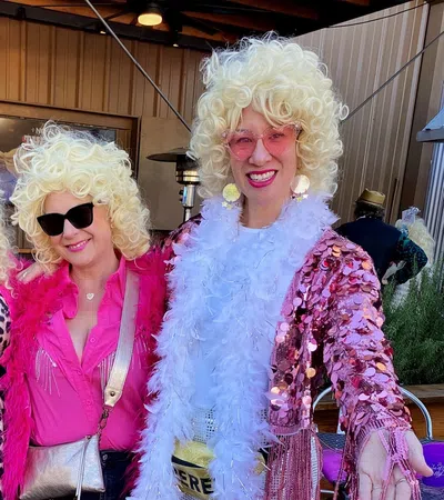 Three women wearing blonde curly wigs and vibrant pink outfits with sunglasses, smiling and posing outdoors.