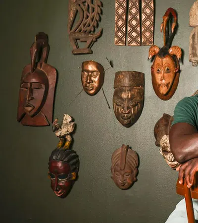 A smiling man poses with African masks hanging on a wall, showcasing cultural art and heritage.