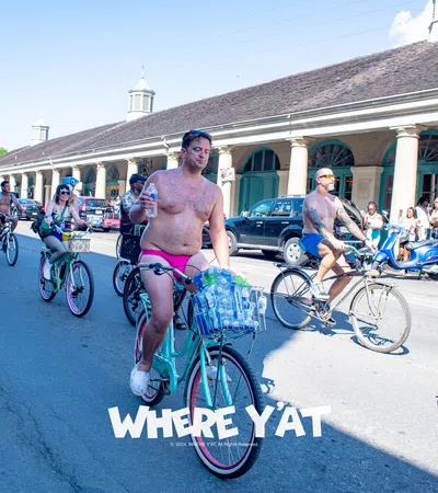Group of minimally clothed cyclists riding on a sunny street during a festive event with spectators watching