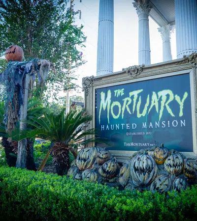 Sign for The Mortuary Haunted Mansion surrounded by Halloween pumpkins and spooky decorations outside a white columned building.