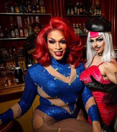 Three drag performers in colorful glamorous outfits pose confidently inside a bar with liquor shelves in the background.