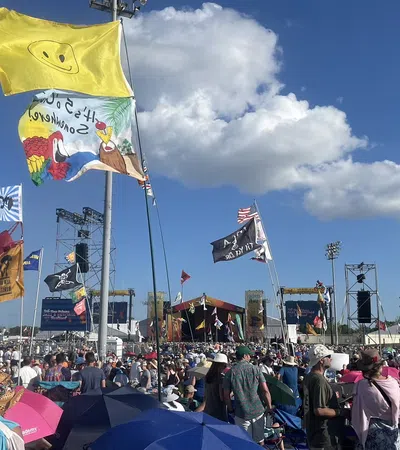 A vibrant festival scene with colorful flags and a large crowd enjoying outdoor music under a blue sky.