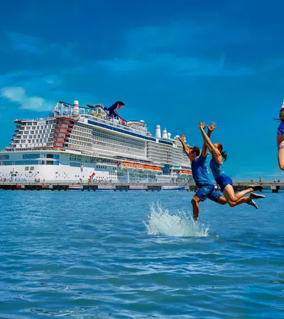Group of children jumping into clear blue water near a large cruise ship docked at a sunny port.